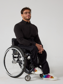  A male model of mixed heritage is looking at the camera. He is wearing black seated twill trousers in regular fit paired with a black shirt and white sneakers. He is sat in a manual wheelchair with his hands resting in his lap.