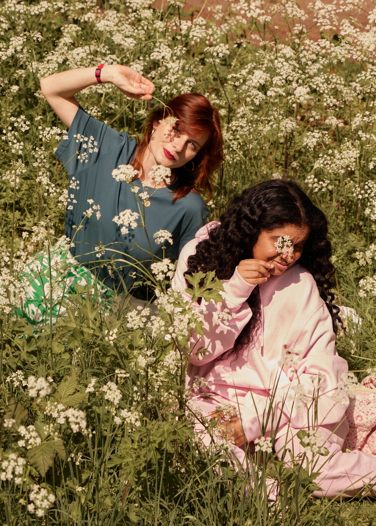Two women sitting in a field of white flowers, surrounded by greenery.