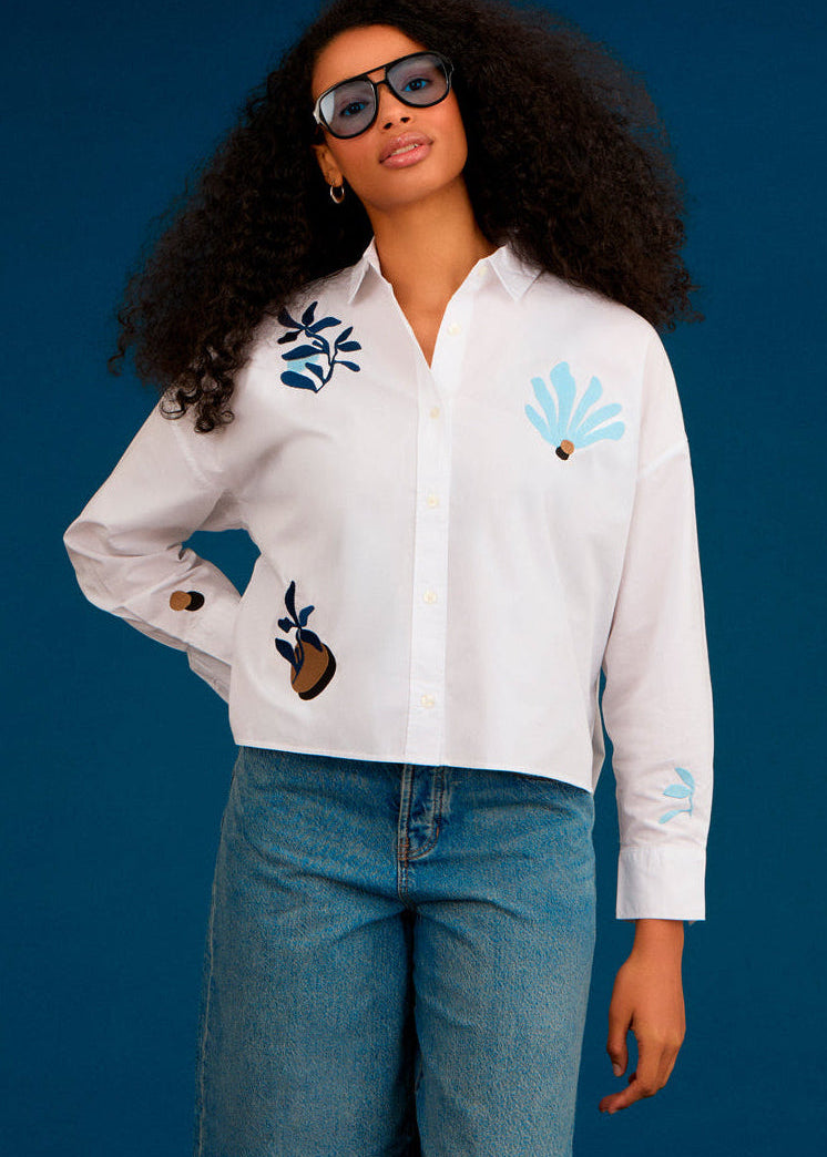 Woman wearing a white shirt with floral designs and denim shorts against a blue background
