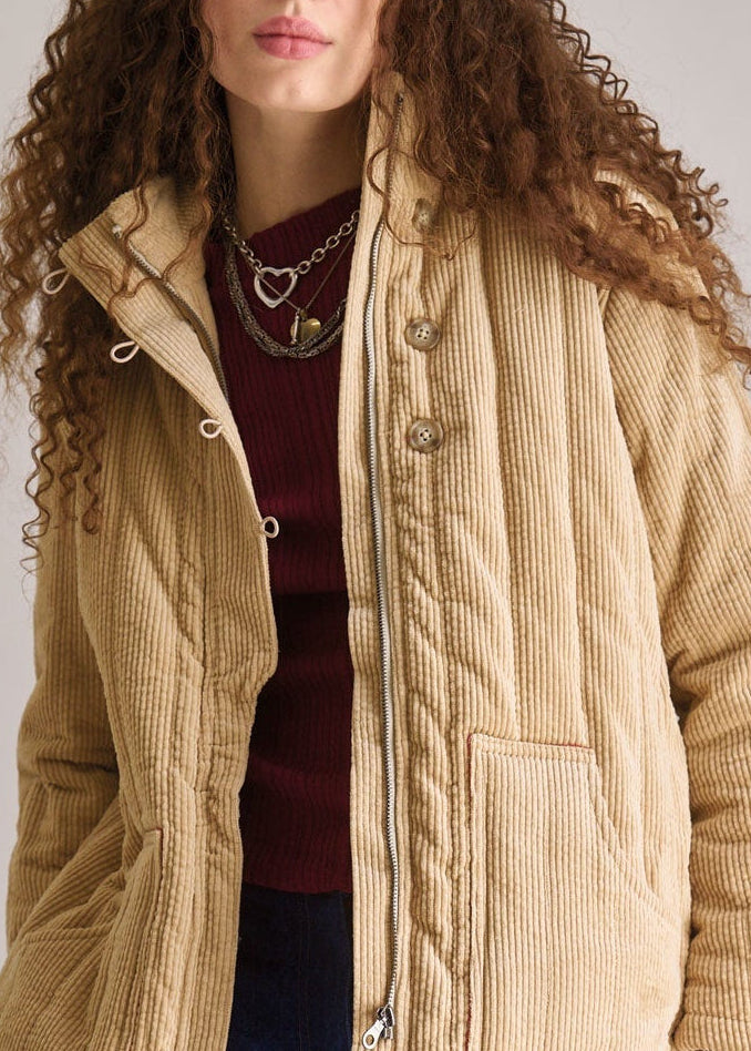 Woman wearing a beige quilted jacket and blue pants against a plain background