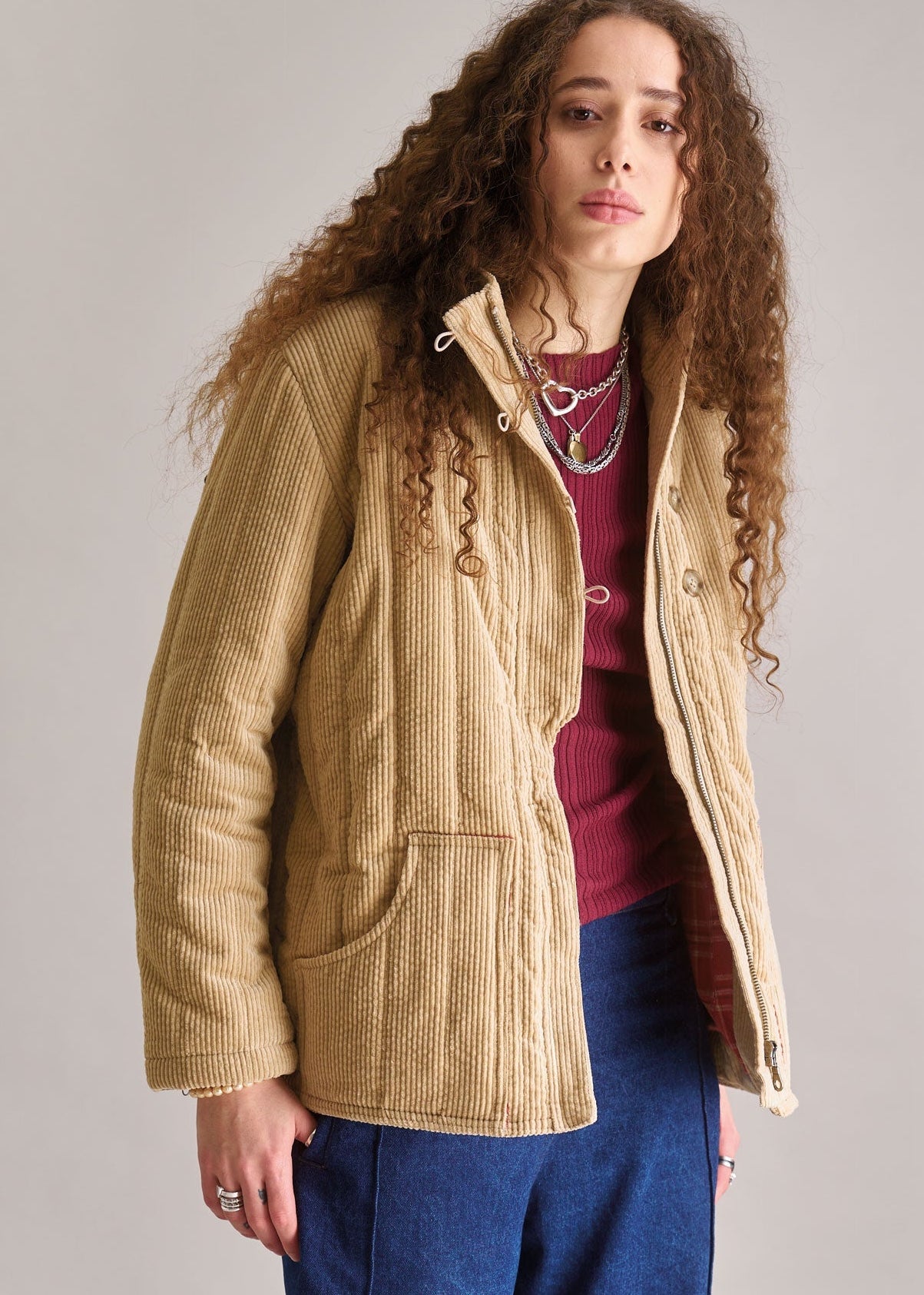 Woman wearing a beige corduroy jacket over a red sweater and blue pants on a plain background