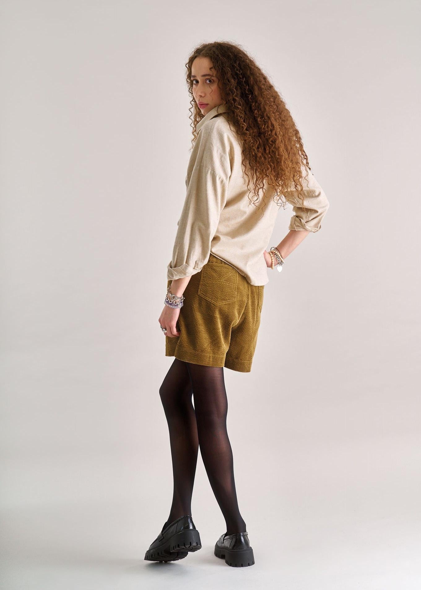 Woman wearing a cream blouse, brown shorts, and black shoes on a plain background