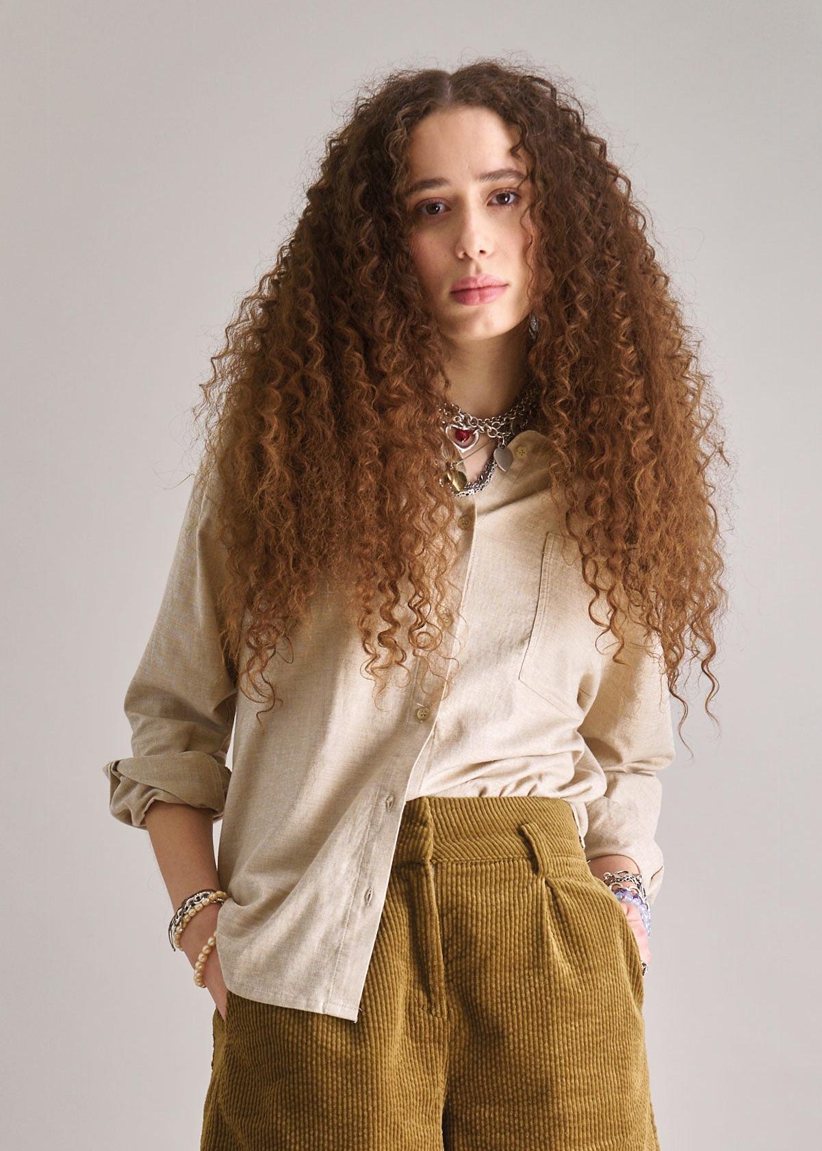 Woman with long curly hair wearing a beige shirt and mustard pants against a plain background