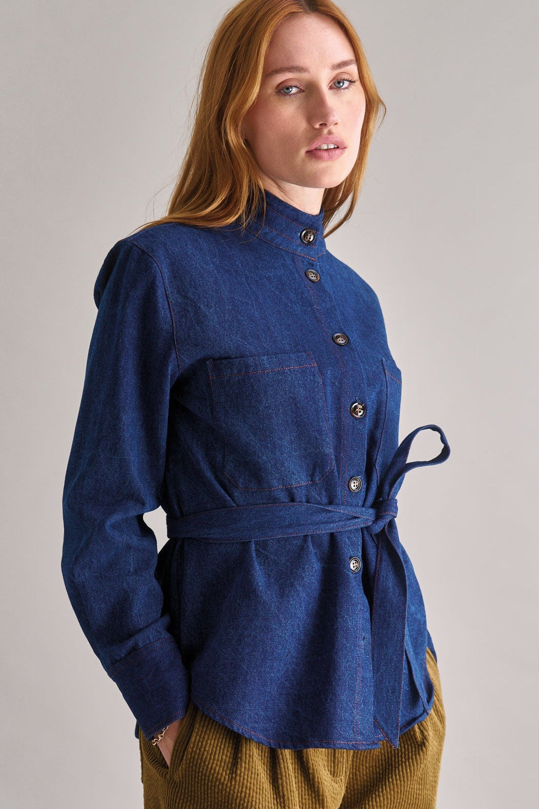Woman wearing a blue jacket with a belt and brown pants on a plain background