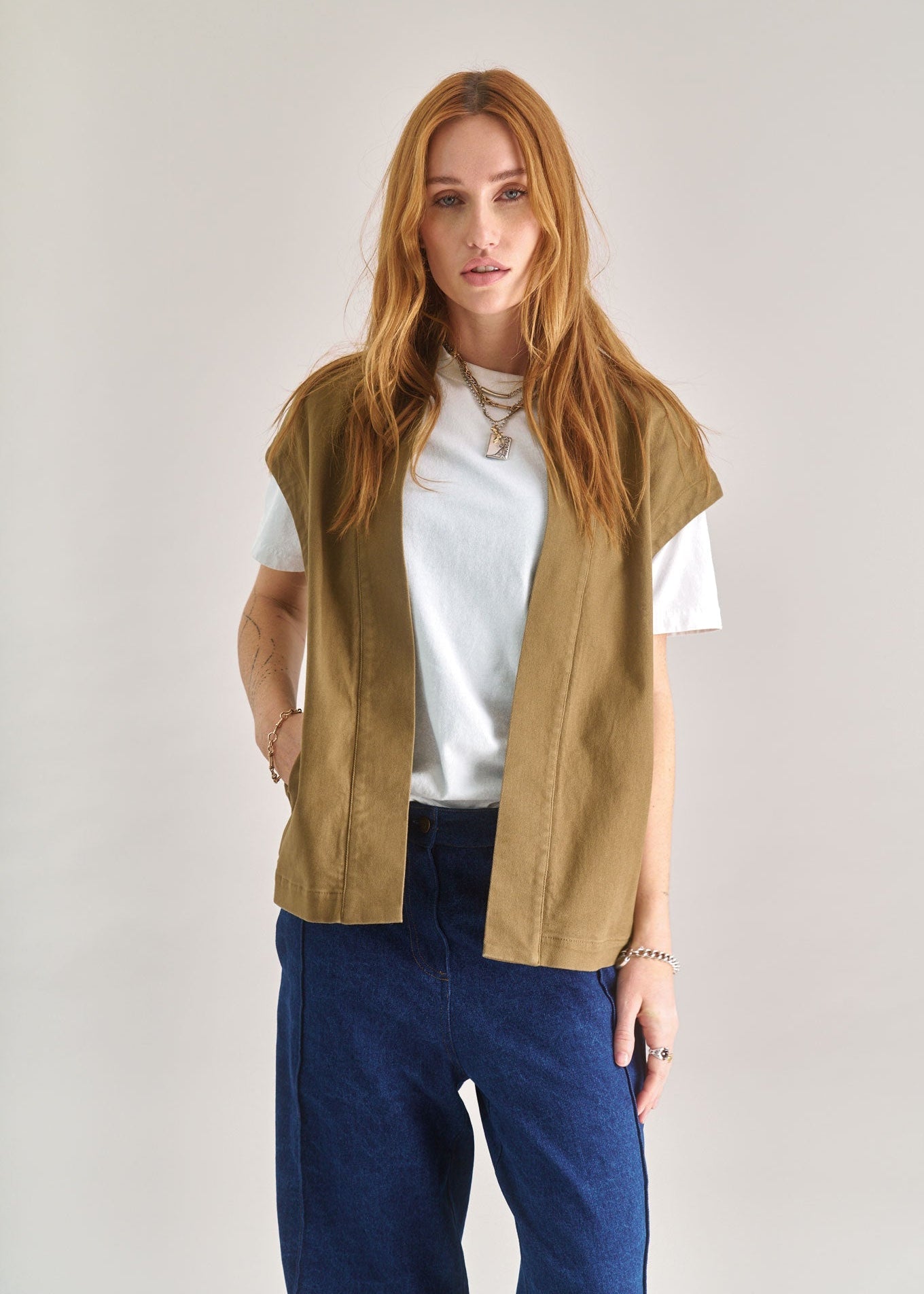 Woman wearing a beige vest over a white shirt and blue pants on a plain background