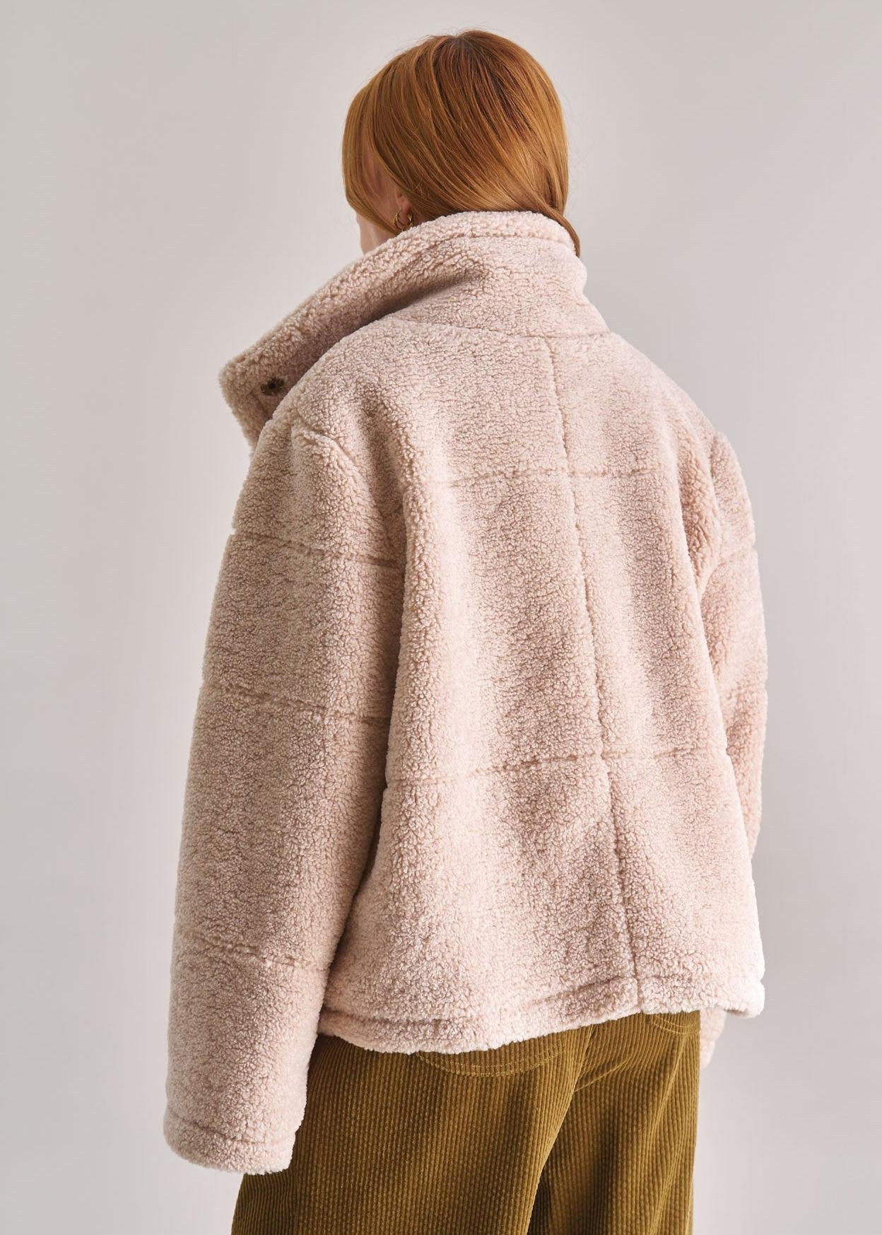 Person wearing a beige fleece jacket and green pants on a light gray background