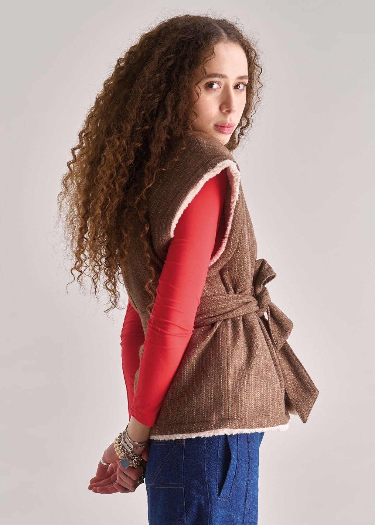 Woman wearing a brown vest over a red shirt with blue pants on a plain background