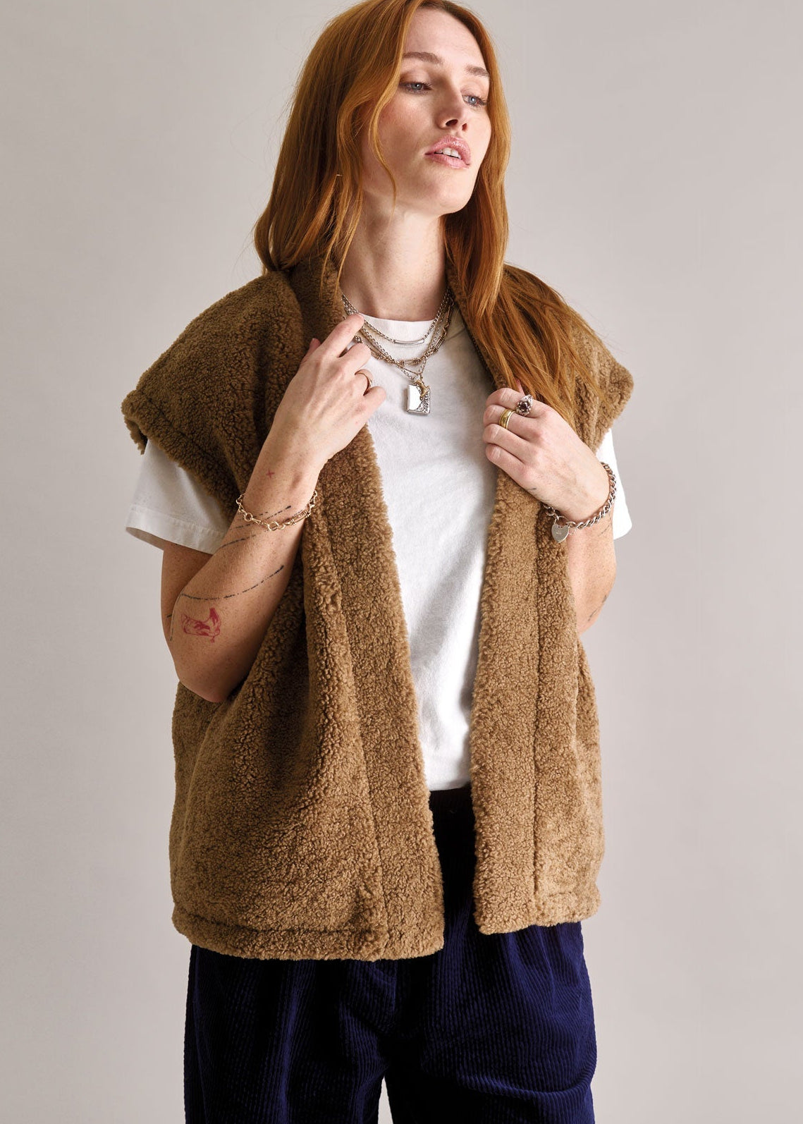 Woman wearing a brown fleece vest over a white shirt and dark pants on a plain background