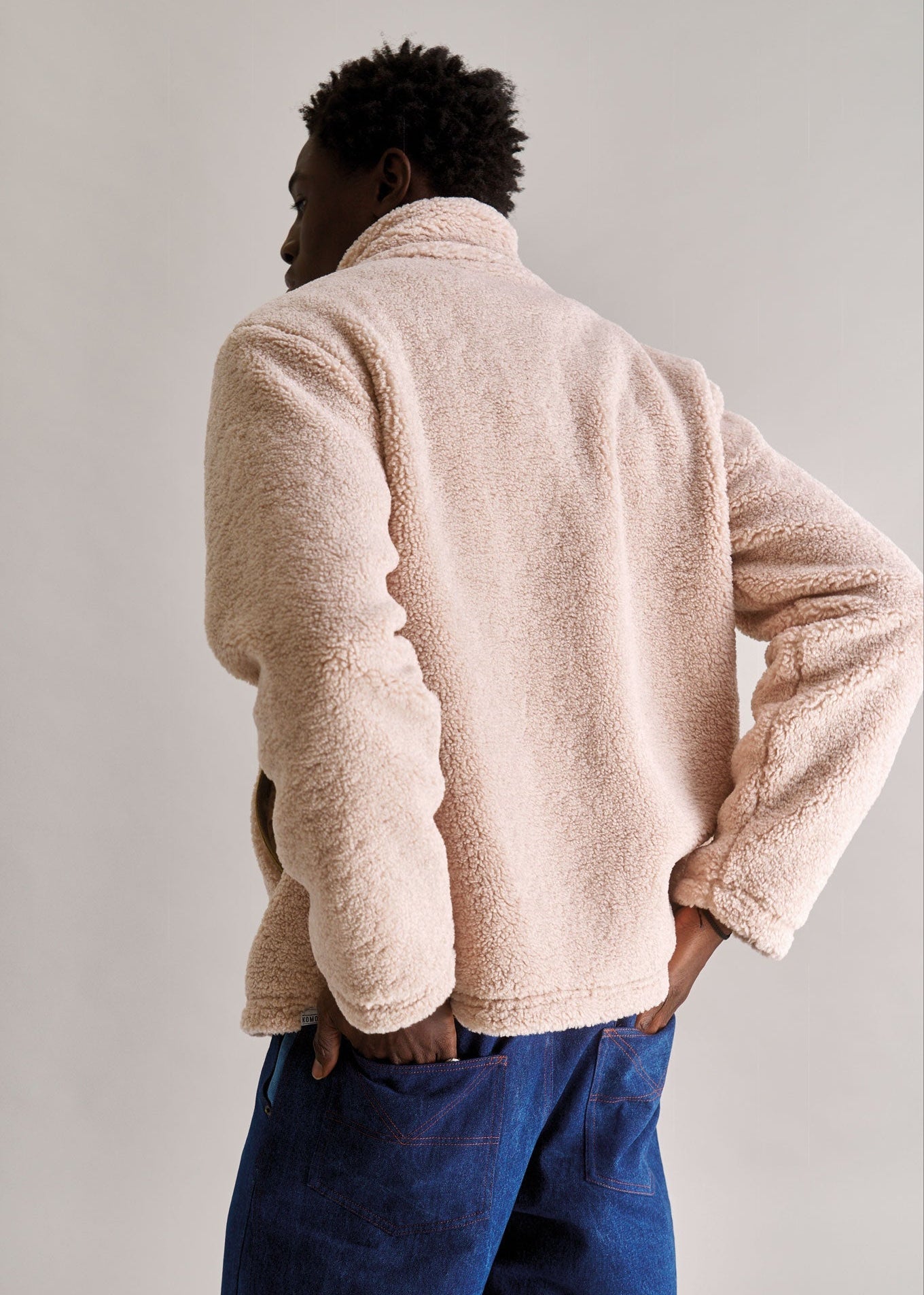 Person wearing a beige fleece jacket and blue pants against a plain background