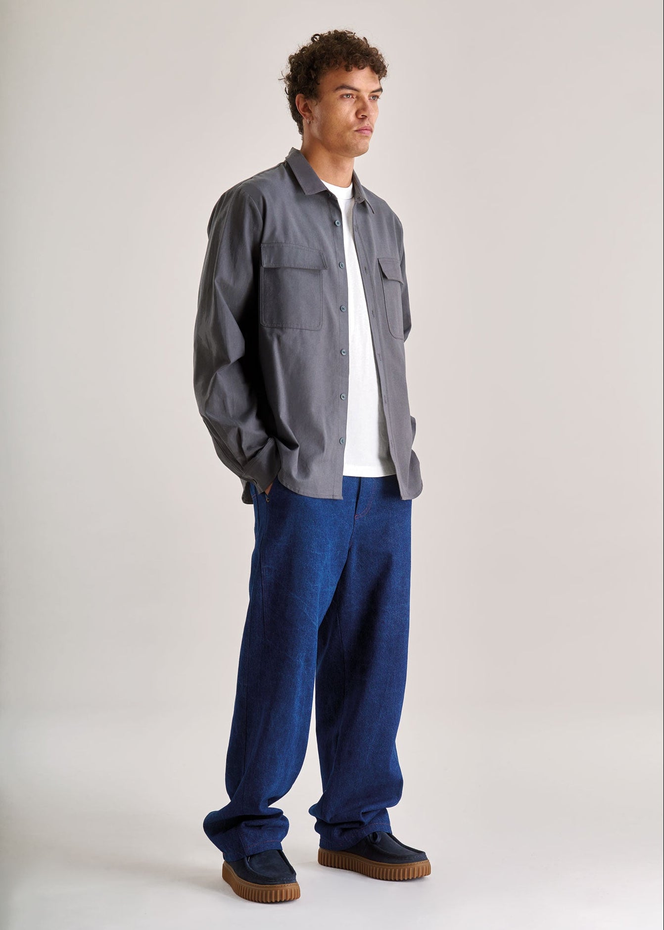Person wearing a gray jacket, white shirt, blue pants, and brown shoes on a light background