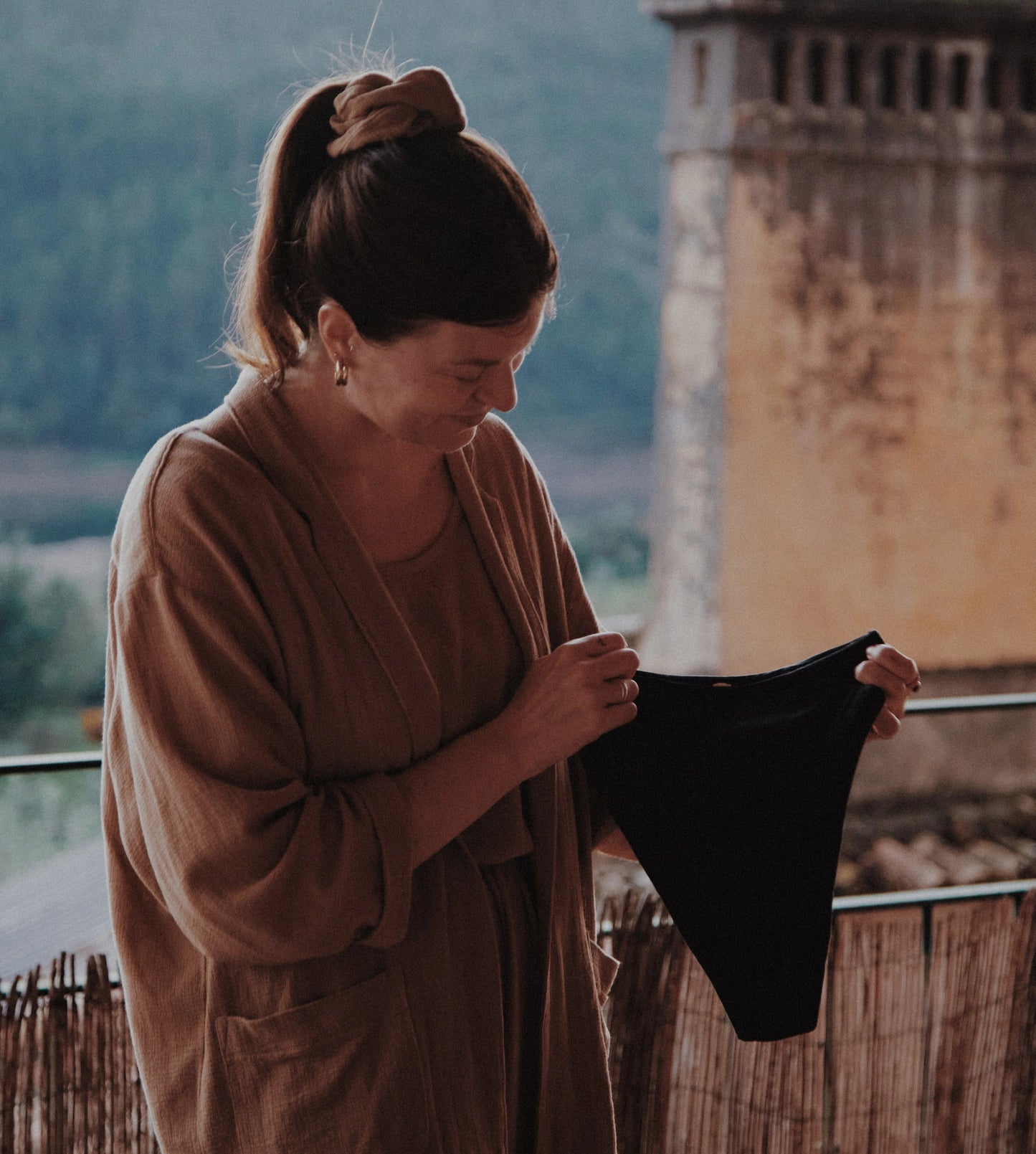 Woman holding a black garment with a scenic background of mountains and an old building.
