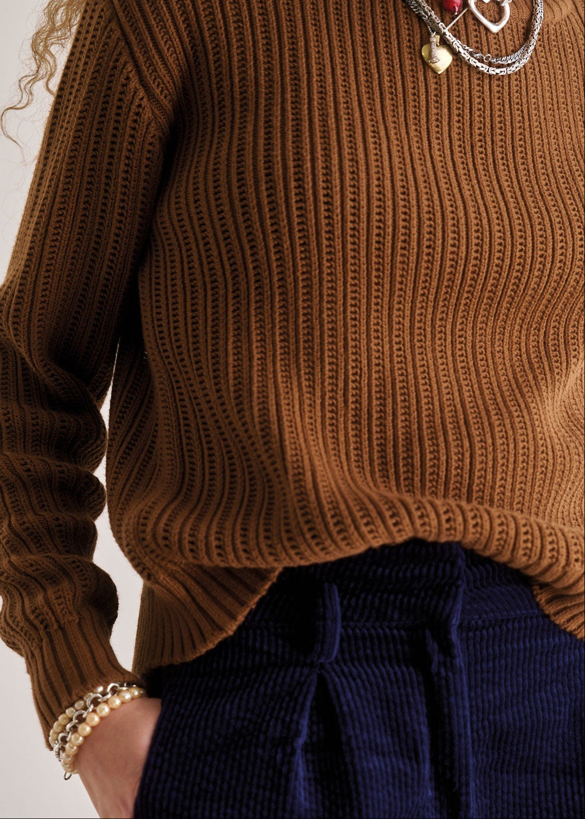 Person wearing a brown ribbed sweater and navy pants with jewelry on a plain background