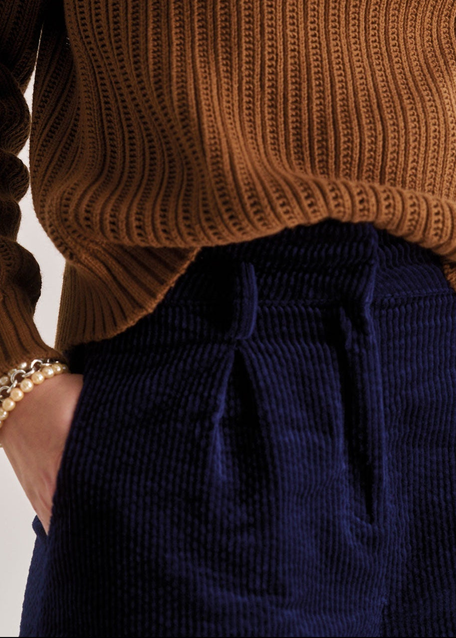 Person wearing a brown ribbed sweater and navy pants with jewelry on a plain background