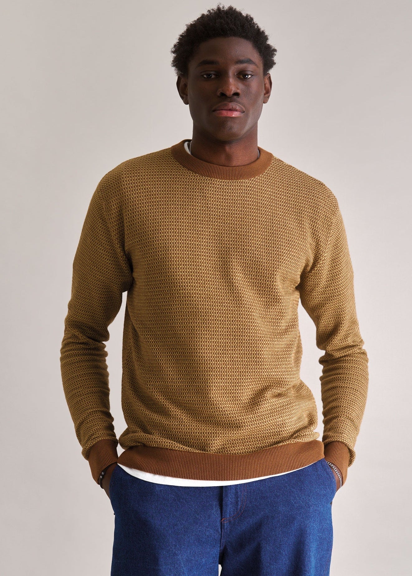 Man wearing a brown textured sweater and blue pants against a plain background