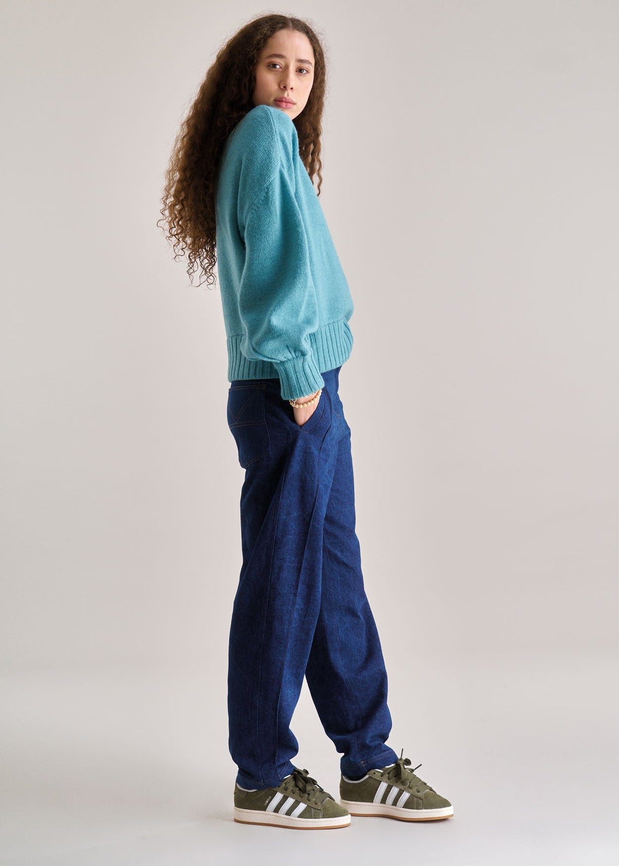 Person wearing a teal sweater and blue pants standing against a plain background