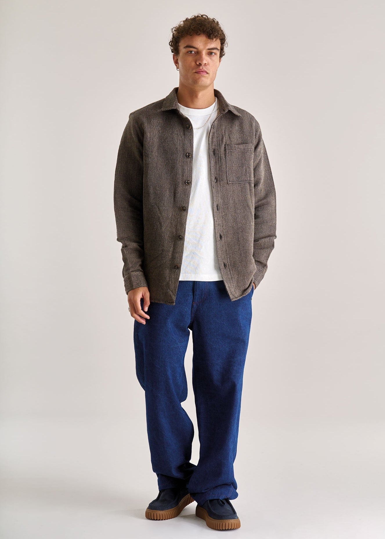 Man wearing a brown jacket, white shirt, blue pants, and brown shoes on a light gray background