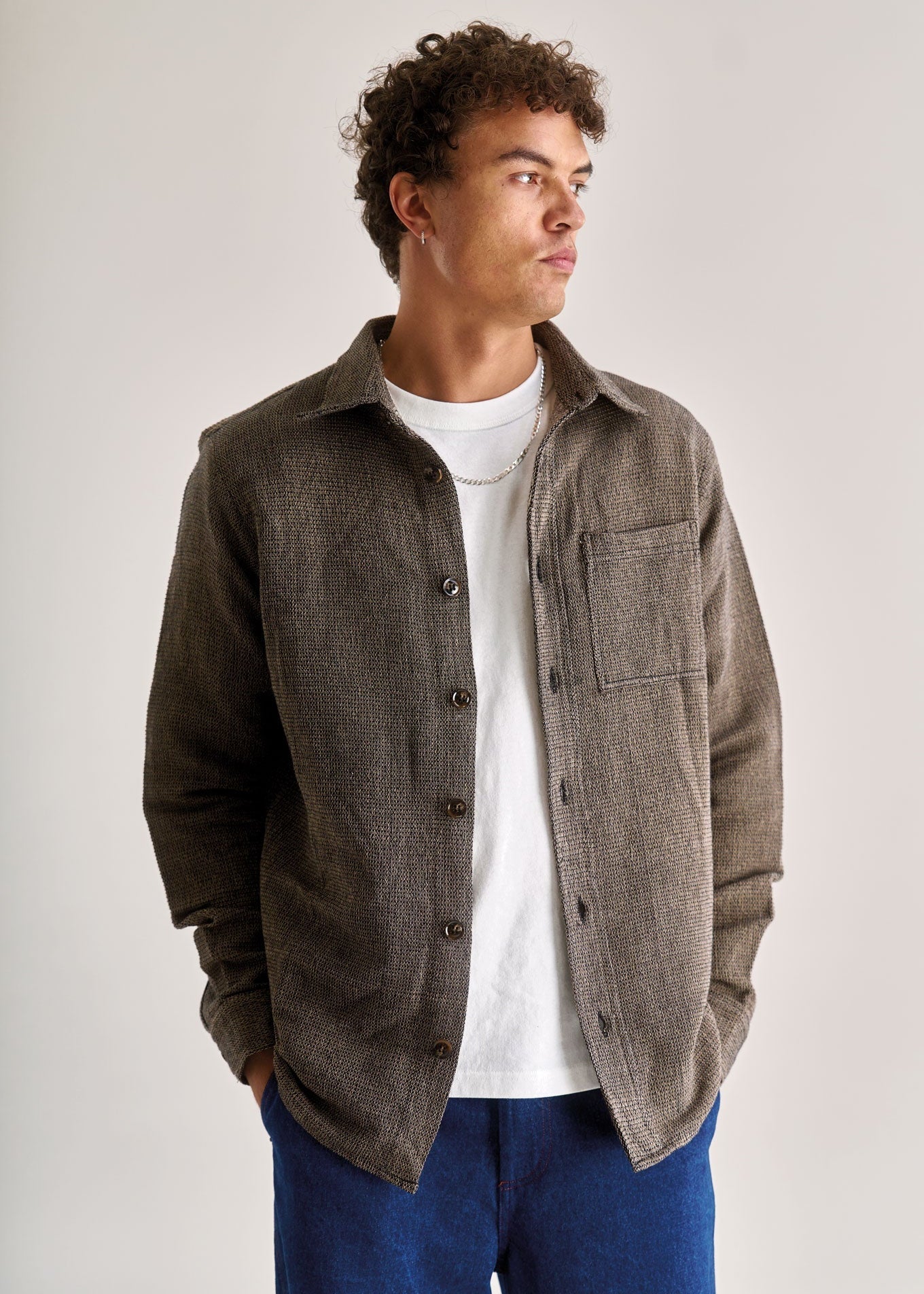 Man wearing a brown jacket over a white shirt and blue pants on a light gray background