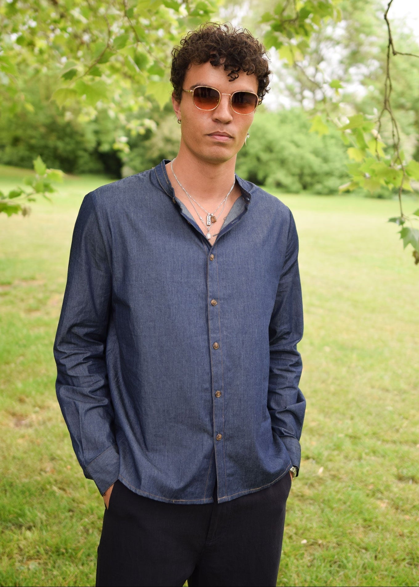 Man wearing sunglasses and a blue shirt standing in a grassy area with trees.