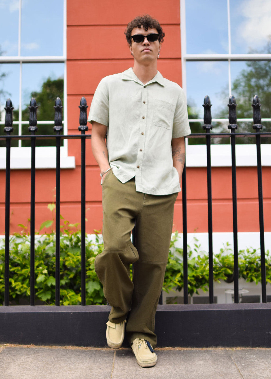 Man wearing sunglasses and a light-colored shirt with dark pants standing in front of a red building.
