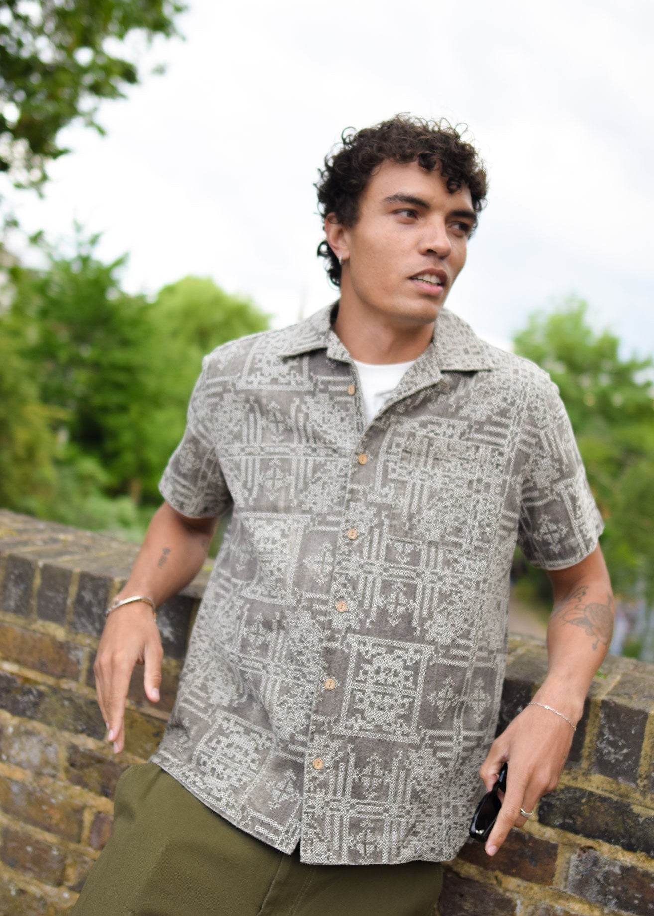 Man wearing a patterned shirt standing outdoors with greenery in the background