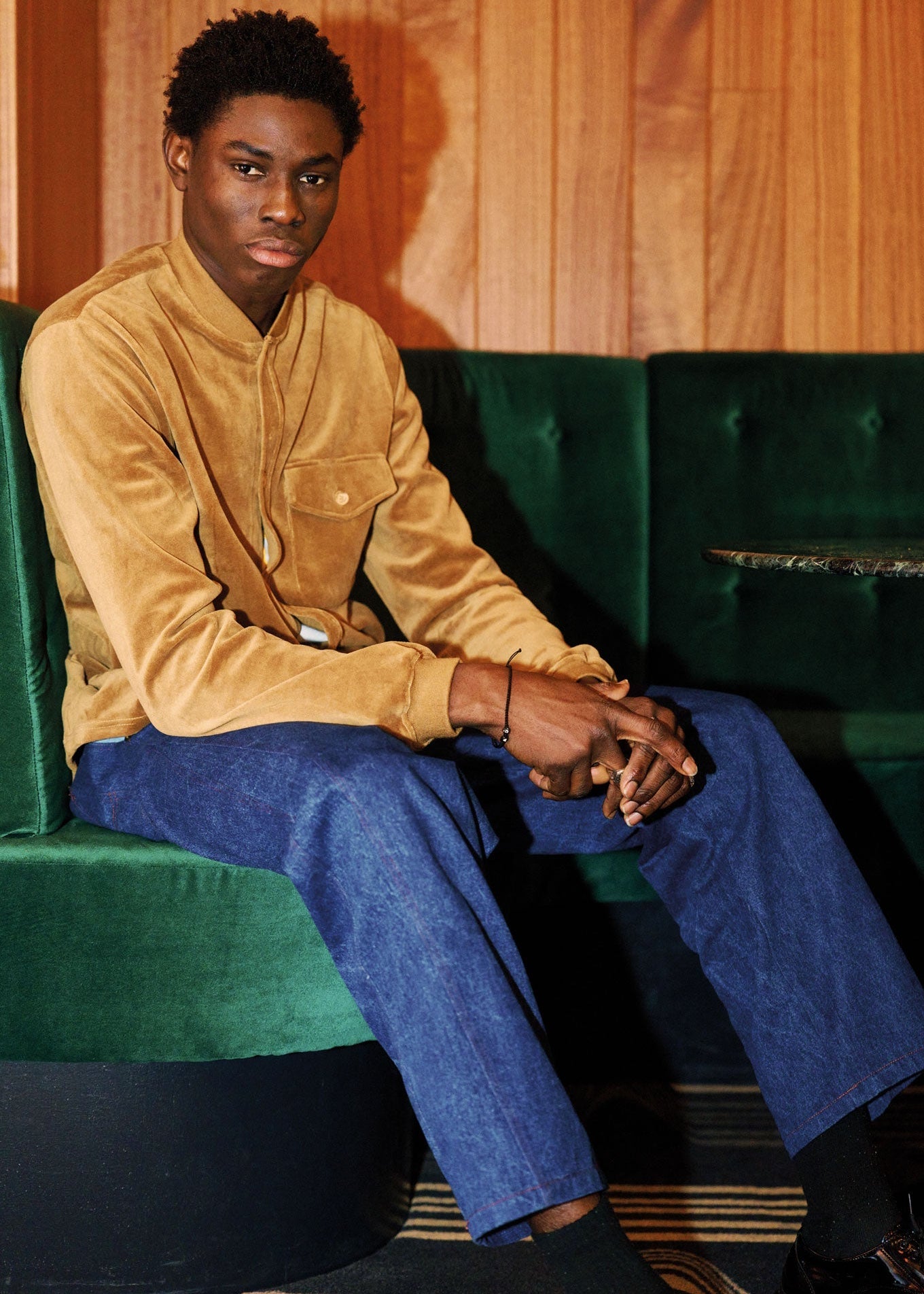 Man sitting on a green couch wearing a mustard jacket and blue pants.