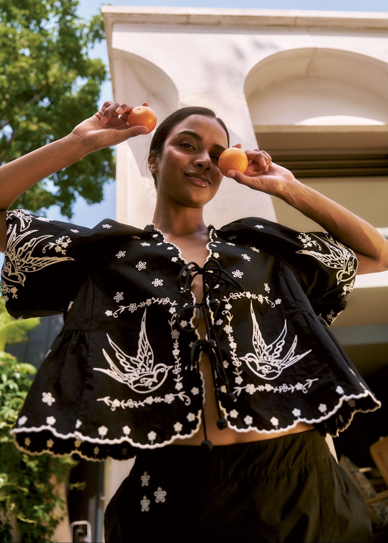 Woman wearing BEATRIX Organic Cotton Top - Black, holding oranges in a sunny outdoor setting.