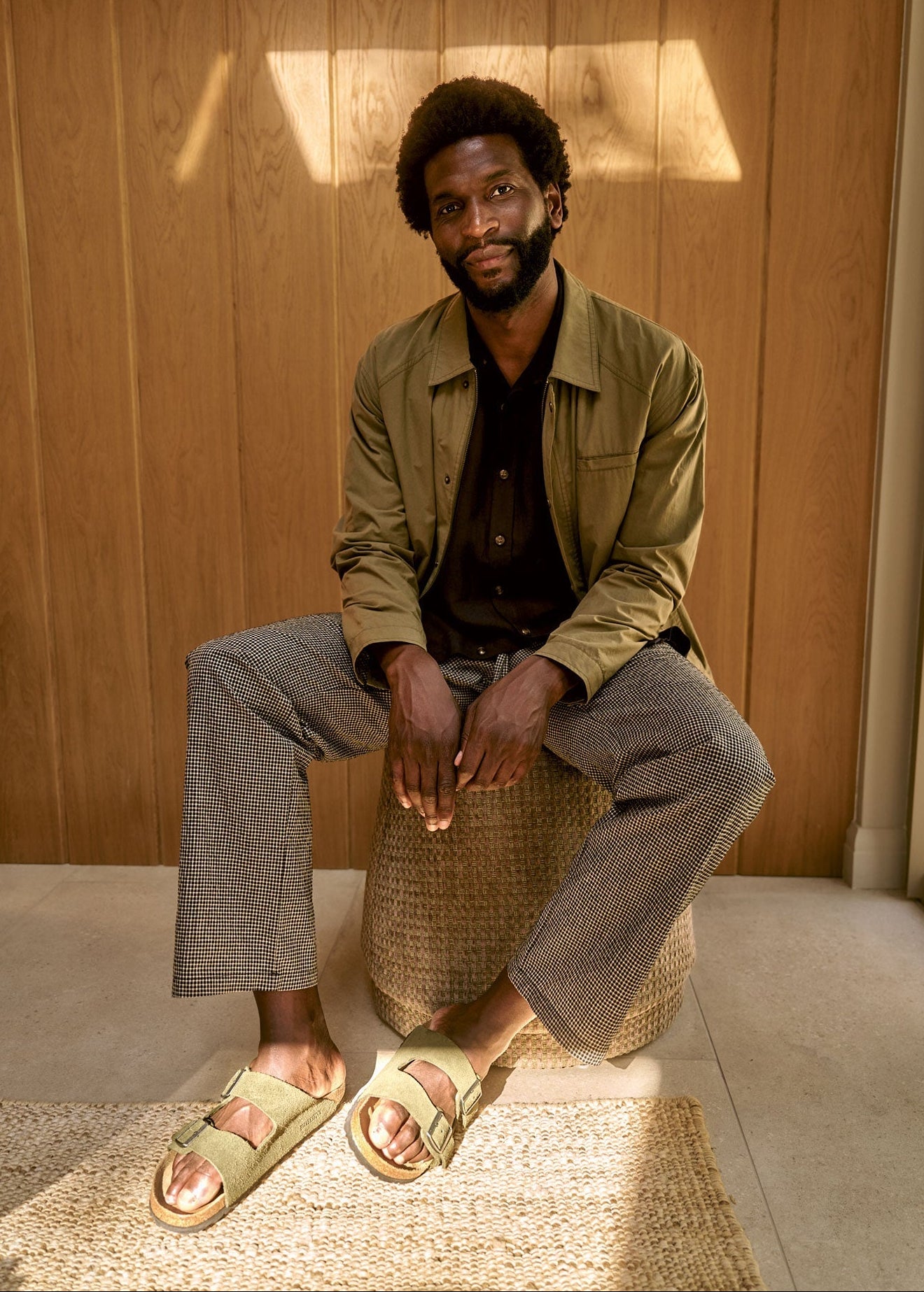 RUDY Organic Cotton Jacket - Olive worn by a man sitting on a stool in a sunlit room, showcasing a casual outfit.