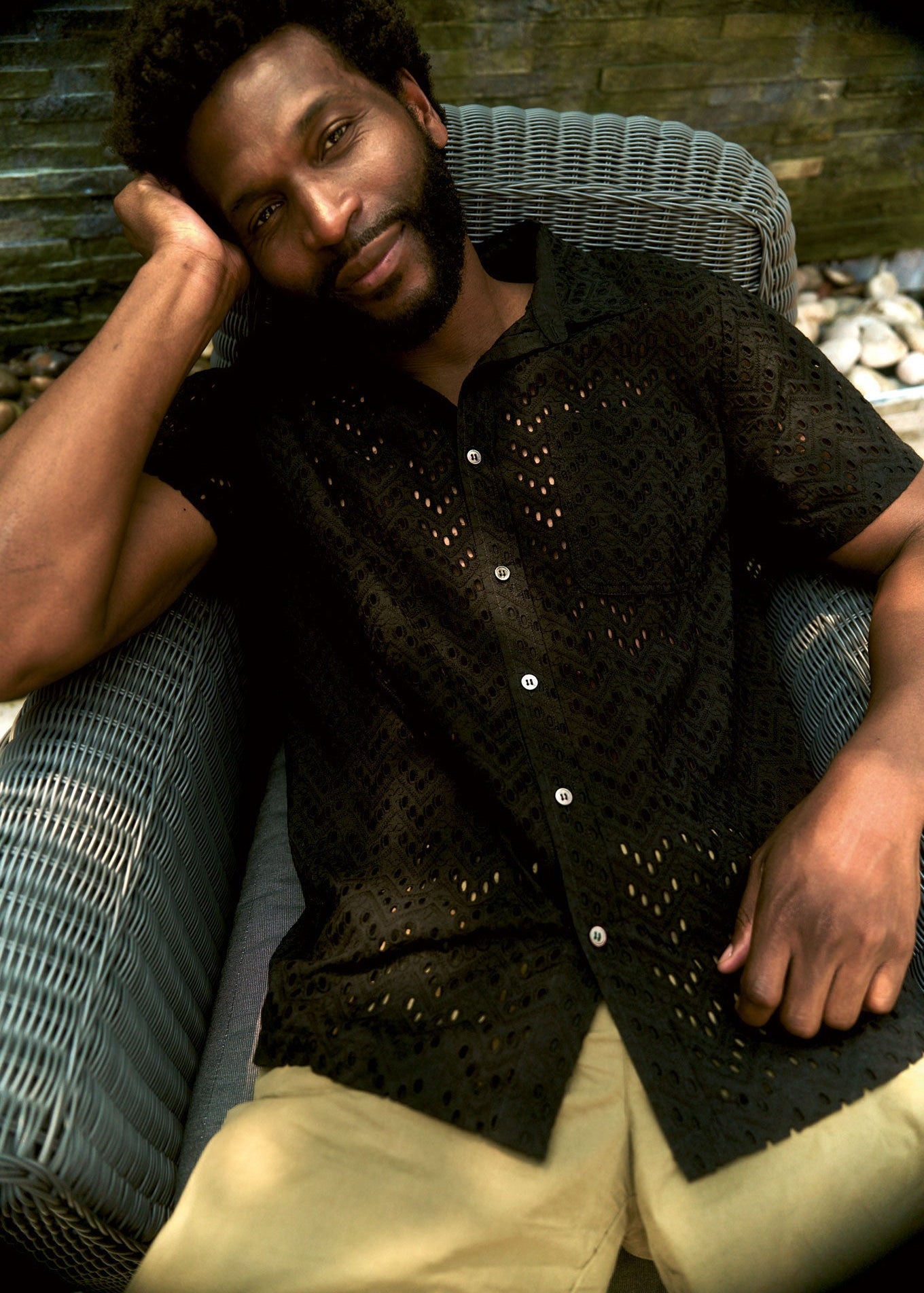 Man sitting on a wicker chair wearing a black patterned shirt with a blurred background