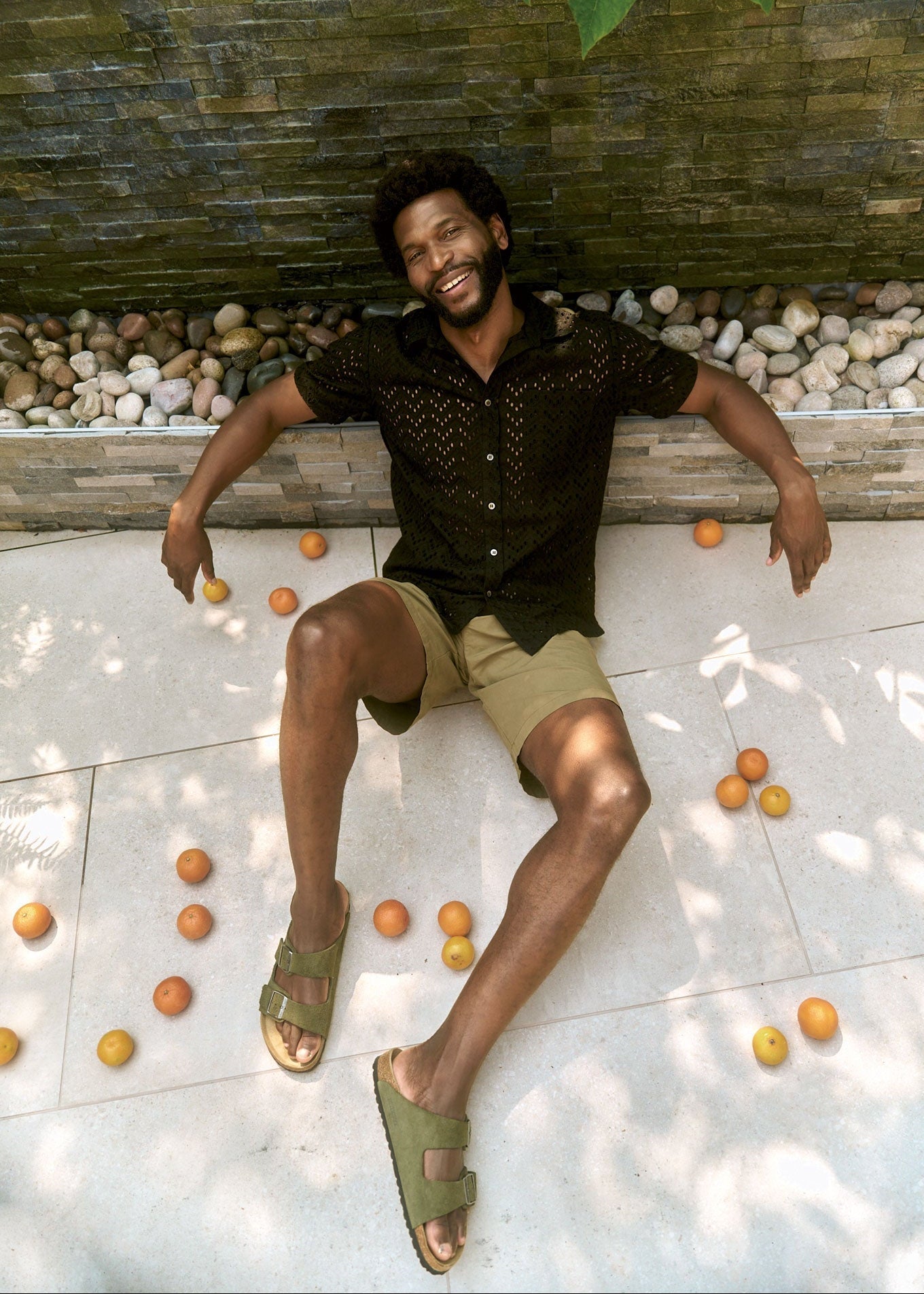 Man lying on a stone floor surrounded by oranges with a stone wall and greenery in the background