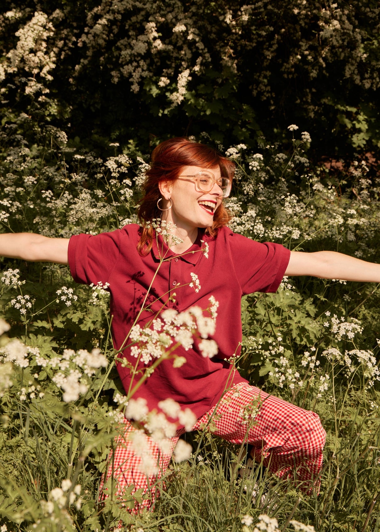 Person in a red shirt and checkered pants standing in a field of flowers with arms outstretched.