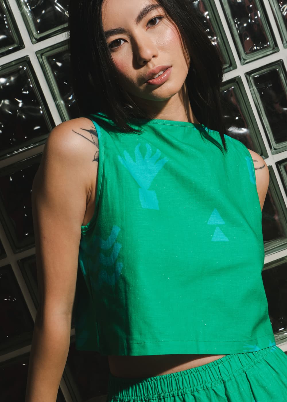 Woman wearing a green sleeveless top with abstract designs against a glass block wall.