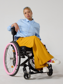  Junior is mixed heritage and has dyed blonde afro hair under a blue silk scarf. They are seated in a wheelchair, wearing a light blue shirt with a collar that has a pocket on the left side as worn. It is cropped at the front. They also have mustard colour jersey culottes on with white sneakers that have sparkly rainbow accents. Set against an off white back ground.