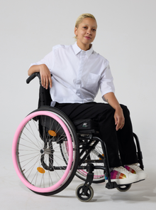  A non-binary model of mixed heritage wears a white short sleeve hi-lo cotton shirt paired with black trousers and shoes with a rainbow patch on them. She is seated in a manual wheelchair with one of her hands resting on the back of the wheelchair while the other hand resting in her lap.
