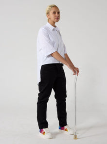  A non-binary model of mixed heritage is standing with a walking stick and looking at the camera, sideways. She is wearing black wrap twill trousers paired with a white shirt and sneakers. Her hands are resting on handle of the clear walking stick. 
