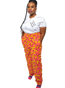  Glynis, a black woman with an arm limb difference, is wearing pink and yellow vibrant , high waisted trousers with a white tshirt with the text slogans "Disabled is not a bad word" .