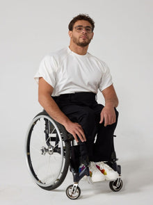  A male model of mixed heritage is looking at the camera. He is wearing black seated 'wrap' twill trousers in regular fit paired with a white t-shirt and white sneakers. He is sat in a manual wheelchair with one of his hands on his knee while the other by his side.