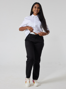  A South asian woman with long back hair is wearing black twill trousers in regular fit paired with a white shirt and sneakers with rainbow patches. She is lifting up her shirt hem to show her stoma bag and the accessibility of the trousers. 