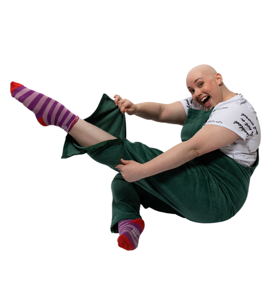 Laura, a white woman with alopecia, is sitting on the floor positioned to the side with one leg pointed out in front of her showing the popper features at the bottom of her trouser leg.  . She is wearing green dungarees with a white t shirt underneath reading "disabled is not a bad word" with stripy purple and pink socks