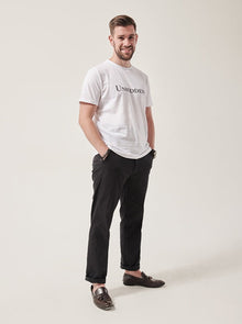  A smiling white man with dark brown hair and dark stubble looks at the camera.  His hands are in his pockets. He is wearing a white t-shirt with Unhidden on it and black trousers. He has brown brogues on.