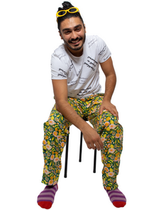  Moeed is a south Asian man, with black hair in bun and yellow sunglasses on his head. He is sitting on a black stool against a white background wearing seated floral yellow and green printed trousers with a white t-shirt with the slogan "Disabled is not a bad word" written across it. He is smiling at the camera.