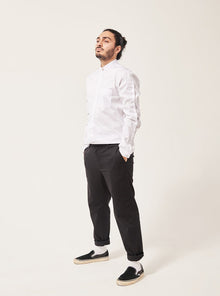  A south Asian man stares moodily off camera. His hair is tied up, his hands are in his pockets. He is wearing a white shirt with a seam down the sleeve and a chest pocket. The collar is a mandarin style with a shell button, and the black trousers are rolled at the hem showing his white socks and plimsoll shoes. The background is tones of grey.