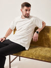 A white man with styled dark brown hair looks to the right. He is reclining on a crushed velvet moss coloured sofa. He is wearing a white t-shirt with wrap sleeves and concealed front zips to allow chest access. His left arm is resting on the back of the sofa and he has a large black watch on.