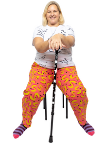  Sylvia, a white woman, is wearing pink and yellow vibrant , high waisted trousers with a white tshirt with the text slogans "Disabled is not a bad word". She is sitting on a black stool and is also wearing purple and pink stripy socks. She is smiling at the camera and holding her arms out in front of her holding a black mobility aide with white stars on it
