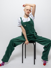 Hannah, a white woman, is sitting on a black stool with her legs apart and her arm resting on her head. She is wearing green dungarees with a white t shirt underneath reading "disabled is not a bad word" with stripy purple and pink socks